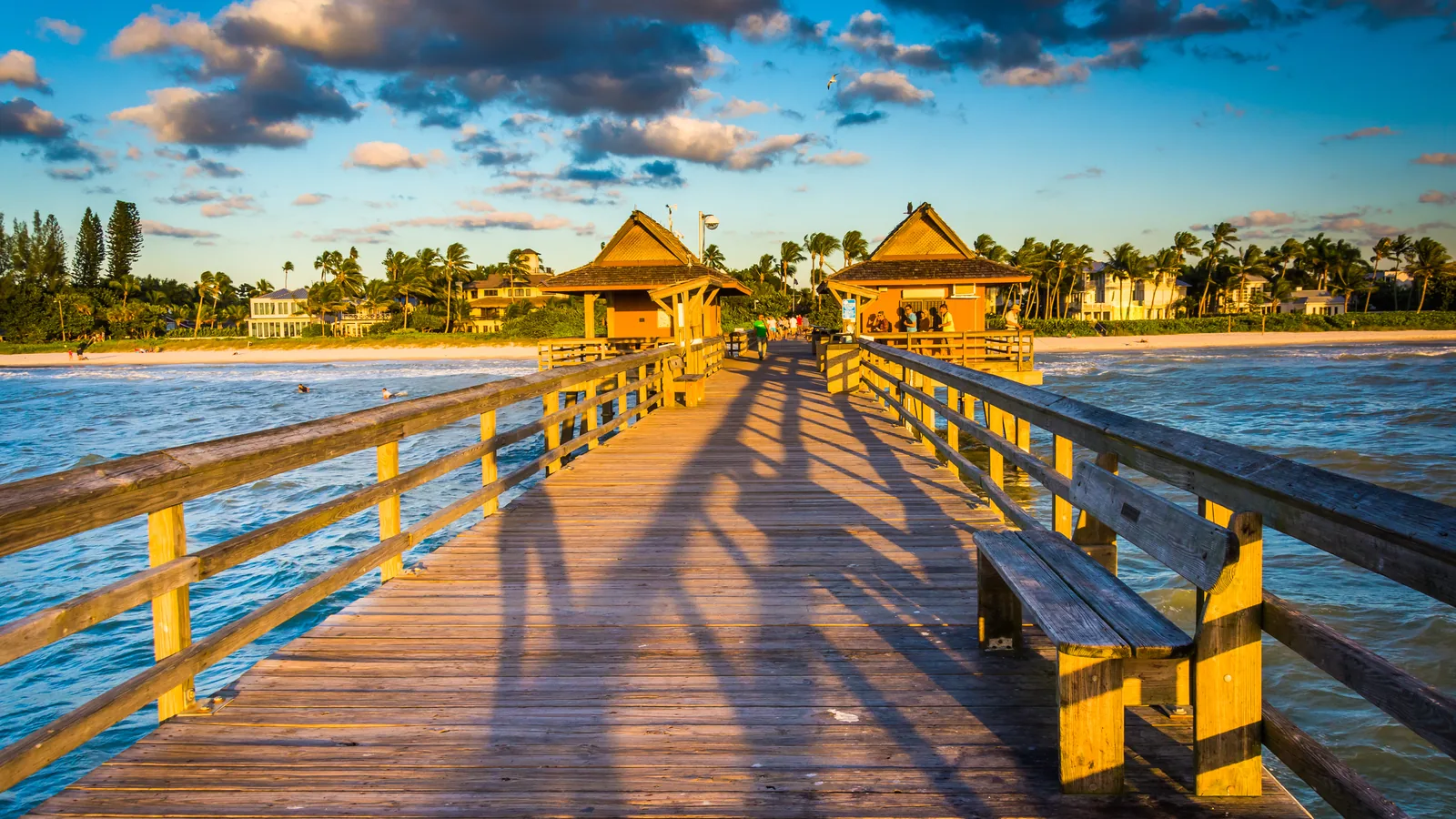 Naples Pier at sunset, a long wooden pier with railings and a bench leading to covered pavilions over the Gulf, with palm trees and beachfront homes along the shore